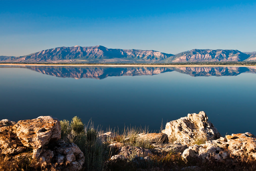 Scenic lake with mineral-rich rocks, clear water, and mountains reflected under blue sky