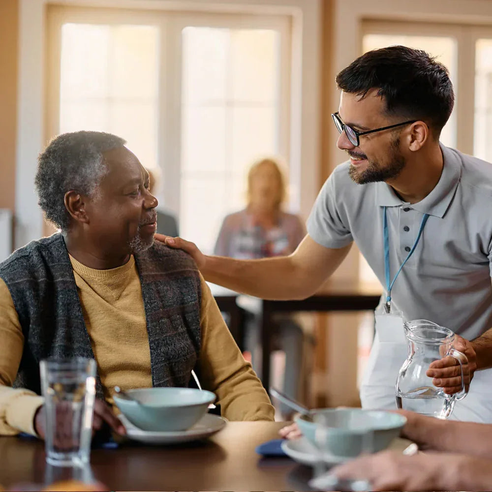 Care worker smiling and supporting elderly man at dining table with water and healthy meal