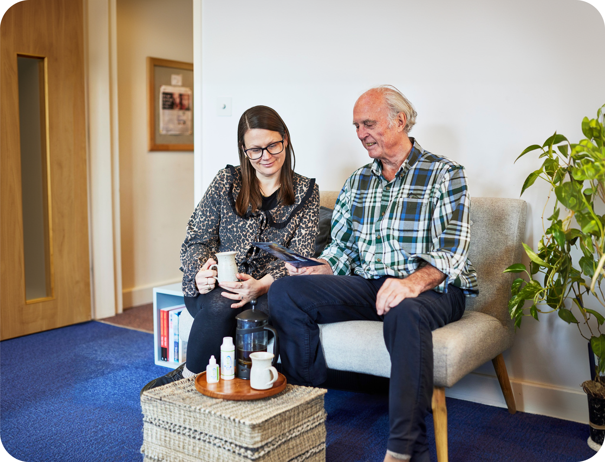 Two people sitting on a couch in a room with a plant and door in the background.