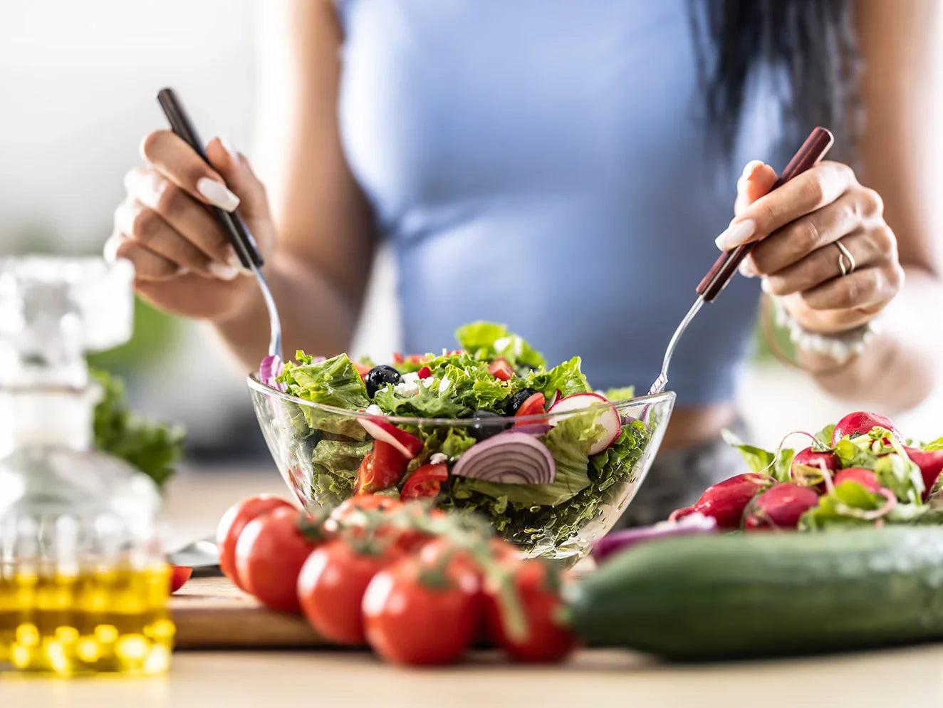 Person holding a fork and knife over a bowl of salad with fresh vegetables on a table.