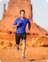Man running in a blue shirt and black shorts against a desert landscape with large rock formations.